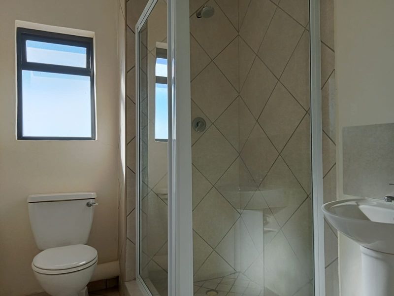 A functional bathroom featuring a shower enclosure, pedestal basin, and toilet, with diamond-patterned wall tiling and a window for natural light.