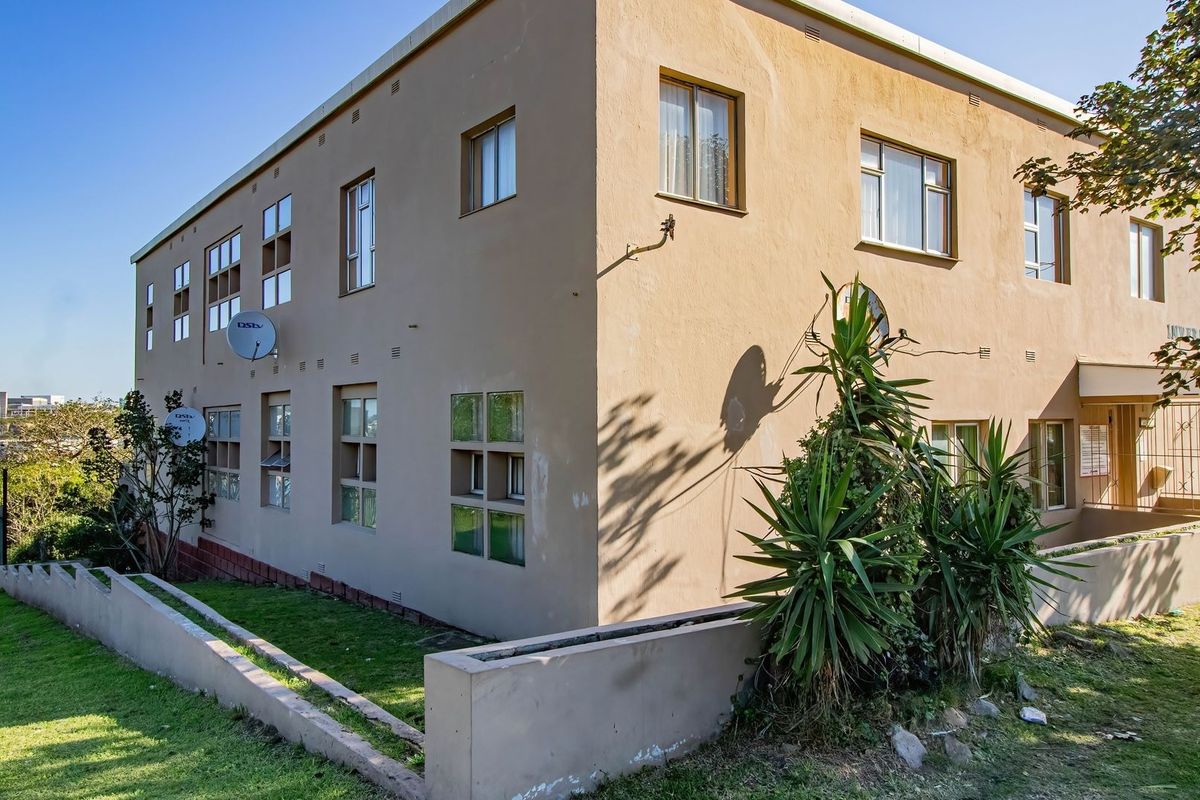 Exterior view of the apartment building featuring a plastered facade, aluminium window frames, and a surrounding retaining wall with landscaped garden area.