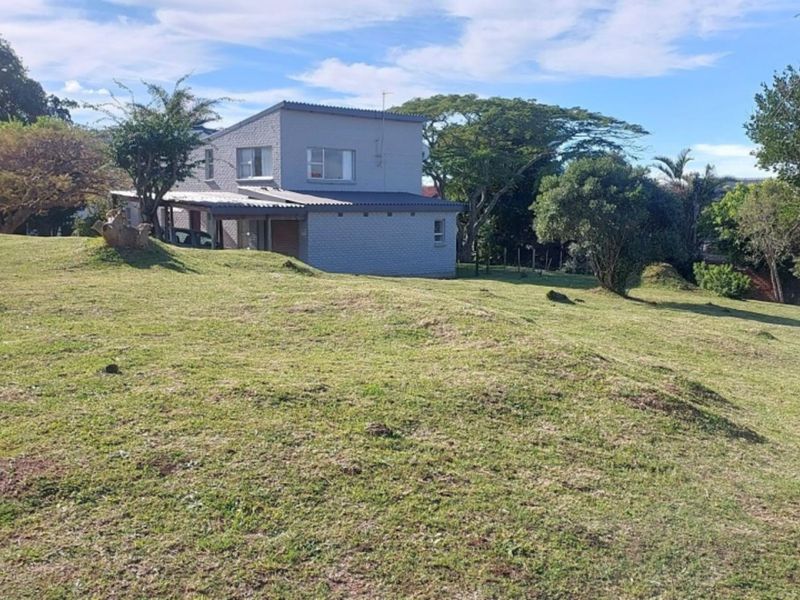 A view of a residential property featuring a sloped grassy lot with mature trees and a paved sidewalk along the street frontage.