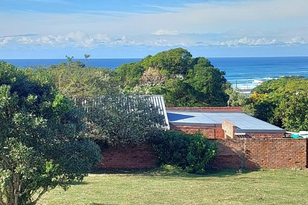 A scenic view overlooking a residential property with a brick boundary wall, mature trees, and a distant ocean horizon.