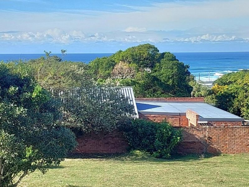 A scenic view overlooking a residential property with a brick boundary wall, mature trees, and a distant ocean horizon.