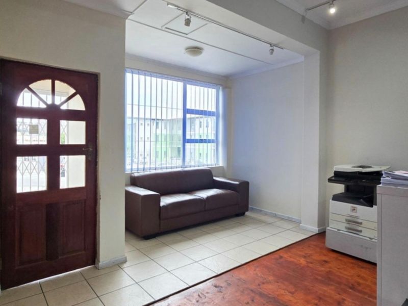 A functional office space featuring a mix of tiled and wooden flooring, track lighting, and a timber entrance door with vertical window blinds.