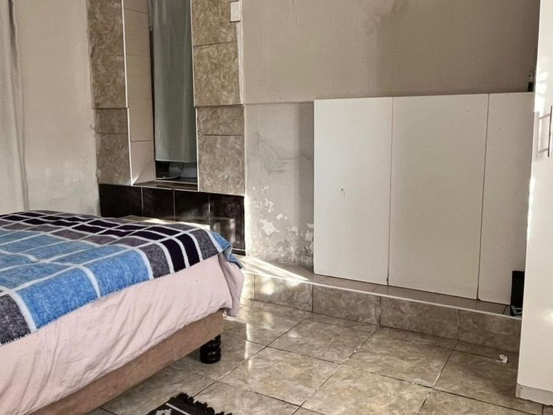 A bedroom featuring tiled flooring and built-in cupboards, showing visible signs of wall surface deterioration and peeling paint requiring maintenance.
