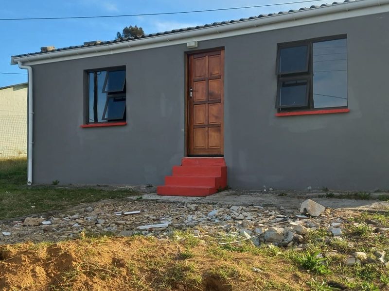 Front exterior view of a single-story house featuring grey rendered walls, timber entrance door, and aluminium windows with unfinished surrounding ground area.