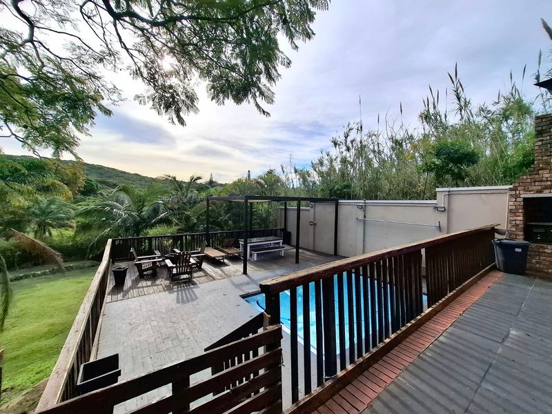Outdoor entertainment area featuring a swimming pool, timber decking, pergola, and a built-in braai, set against a natural landscape backdrop.