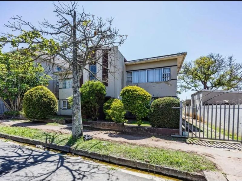 Front exterior view of an apartment building featuring a concrete facade, metal perimeter fencing, and mature landscaping in need of maintenance.