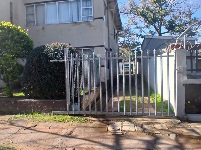 Front exterior view of an apartment building featuring a metal security gate, perimeter wall with razor wire, and a paved driveway.