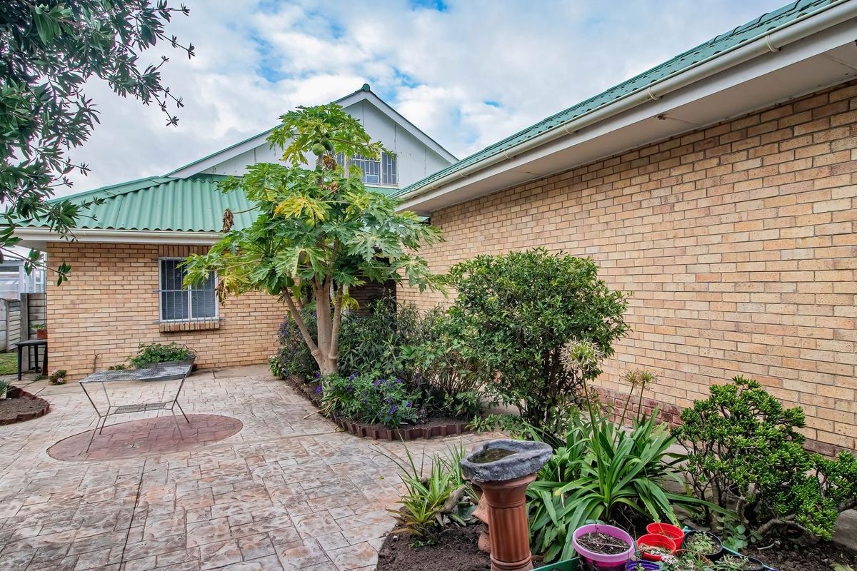A rear exterior view of the house featuring a brick facade, green metal roofing, and a paved patio area with established garden beds.