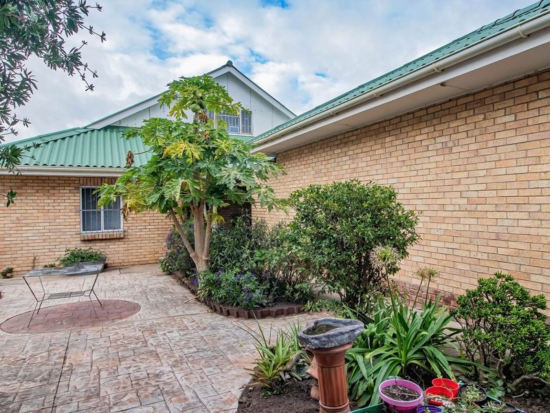 A rear exterior view of the house featuring a brick facade, green metal roofing, and a paved patio area with established garden beds.