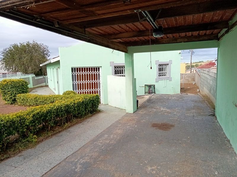 Rear exterior view featuring a covered carport, concrete driveway, and security-fitted windows and doors, set against a boundary wall and garden area.