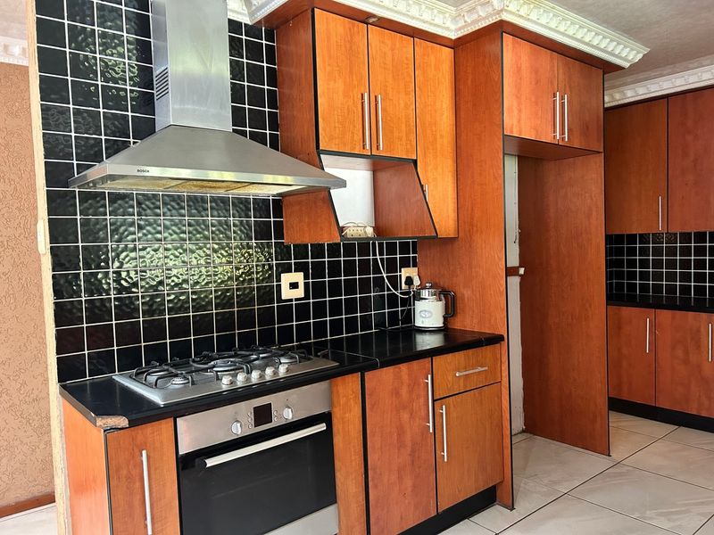 A functional kitchen featuring wooden cabinetry, a gas hob with extractor fan, and a black tiled backsplash with contrasting light floor tiles.