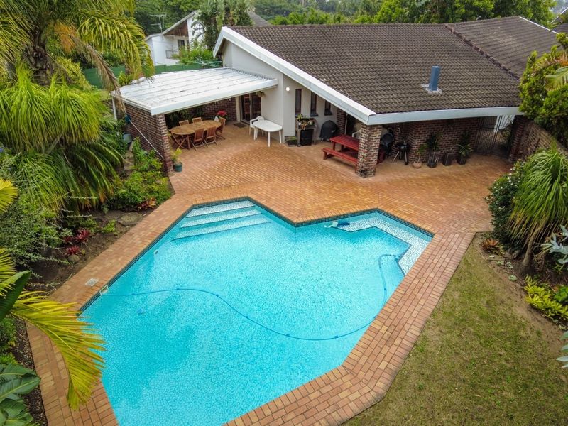 Aerial view of a residential property featuring a swimming pool, brick-paved patio area, covered outdoor space, and established garden surroundings.