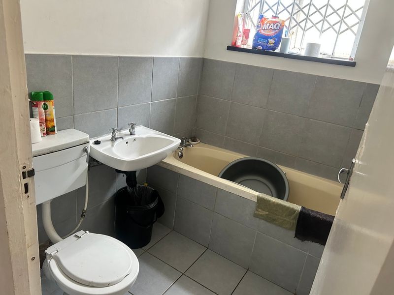 A functional bathroom featuring a pedestal basin, toilet, and bathtub with grey wall tiling and security bars on the window.