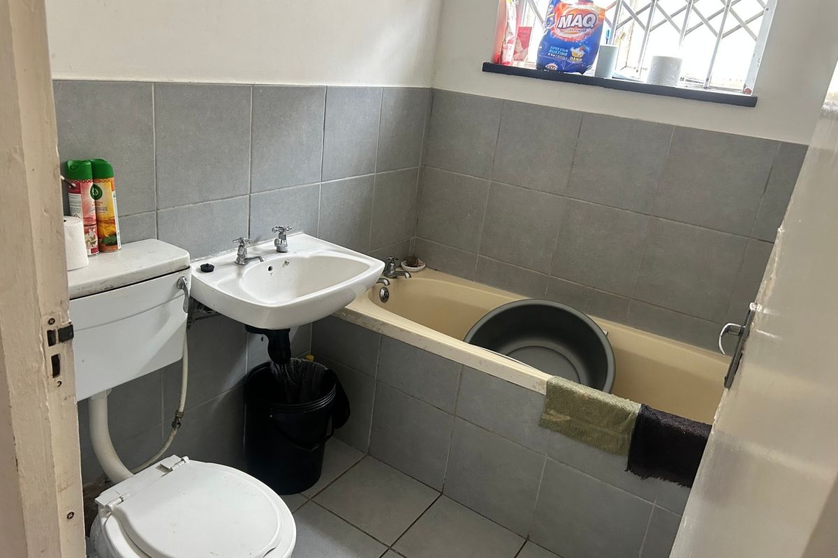 A functional bathroom featuring a pedestal basin, toilet, and bathtub with grey wall tiling and security bars on the window.