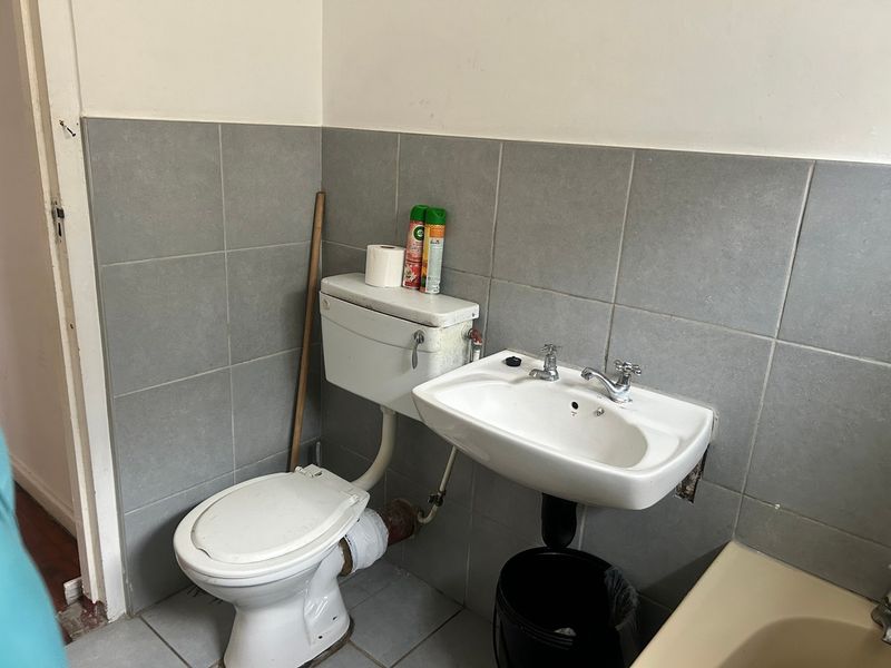 A functional bathroom featuring a wall-mounted basin and standard toilet with grey tiled walls and flooring, currently requiring cosmetic updates and maintenance.