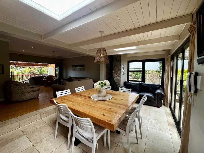 Spacious open-plan living and dining area featuring exposed beam ceilings, skylights, a stone fireplace, and seamless indoor-outdoor flow through large sliding glass doors.