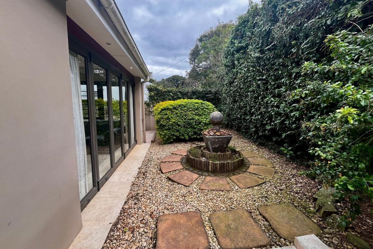 A well-maintained rear garden area featuring stone pavers, a decorative fountain, and established hedging, accessible via large sliding glass doors.