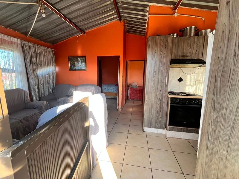 Open-plan living area featuring a compact kitchen with built-in cabinetry, oven, and hob, finished with tiled flooring and an exposed corrugated metal ceiling.