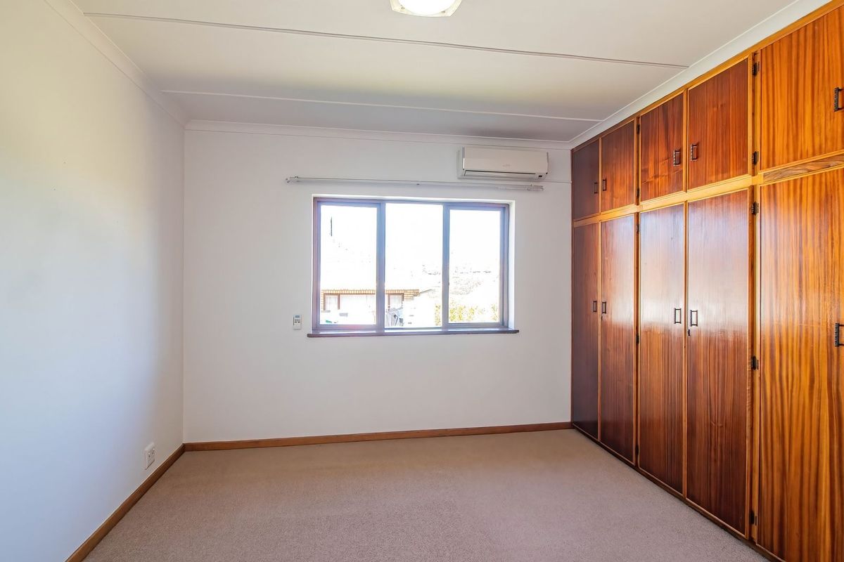 A spacious, empty bedroom featuring wall-to-wall built-in wooden cupboards, carpeted flooring, and a wall-mounted air conditioning unit.