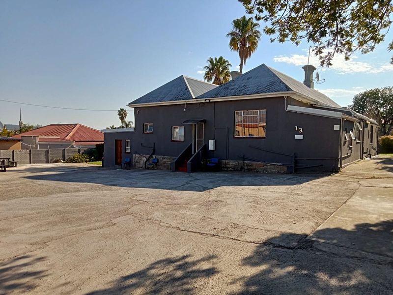 Rear view of the guest house featuring a dark grey facade, corrugated metal roof, and a large concrete parking area with visible surface cracking.
