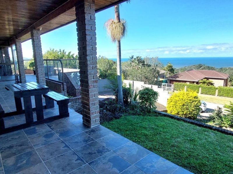 A spacious covered patio featuring tiled flooring, exposed brick pillars, and a timber ceiling, offering scenic views of the garden and distant ocean.