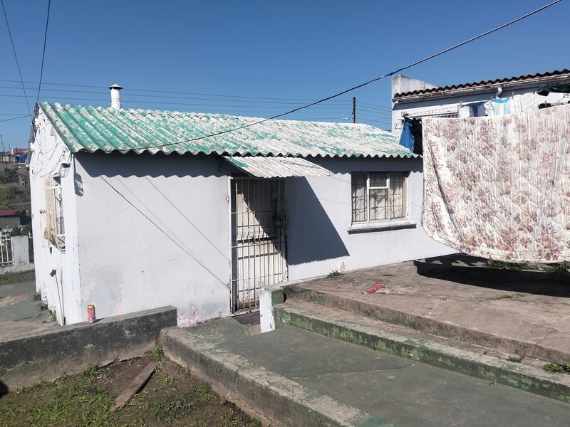 Rear view of a small house featuring a corrugated roof, security gates, and a concrete patio area requiring maintenance.