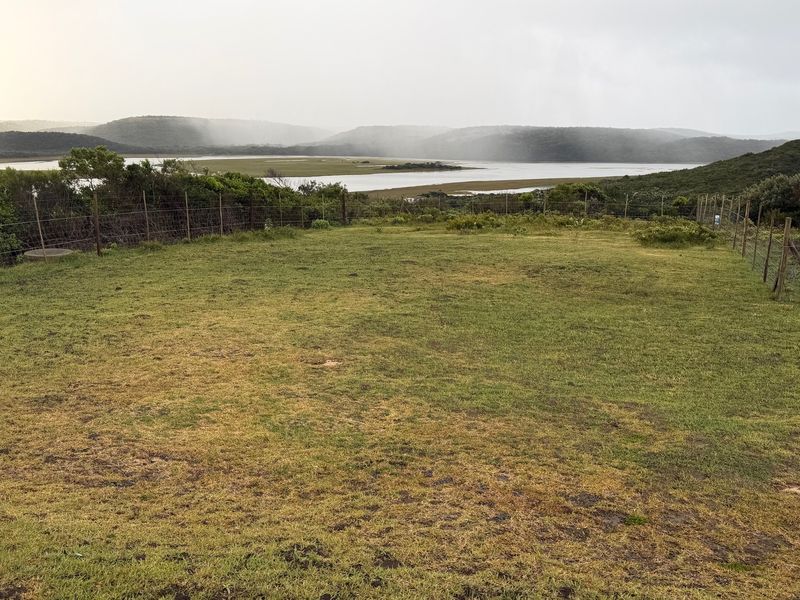 A scenic view of a natural landscape featuring a grassy foreground, wire fencing, and a distant river or estuary under an overcast sky.