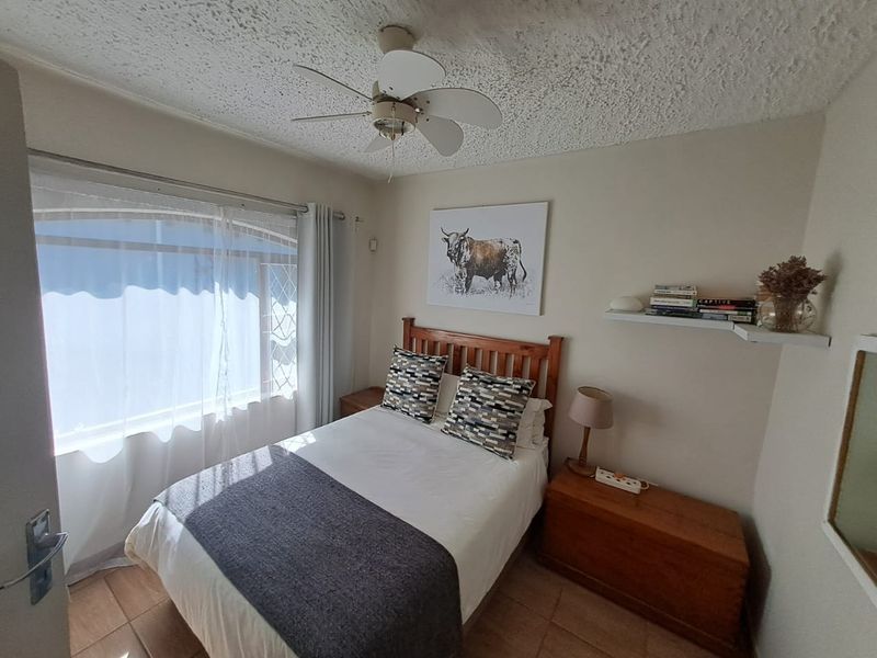 A compact bedroom featuring tiled flooring, a ceiling fan, and a window with security bars, offering a functional and well-lit sleeping space.