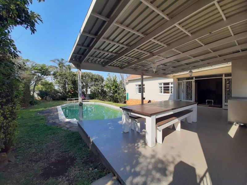 A covered outdoor patio area featuring tiled flooring and a metal roof, overlooking a private swimming pool and garden.