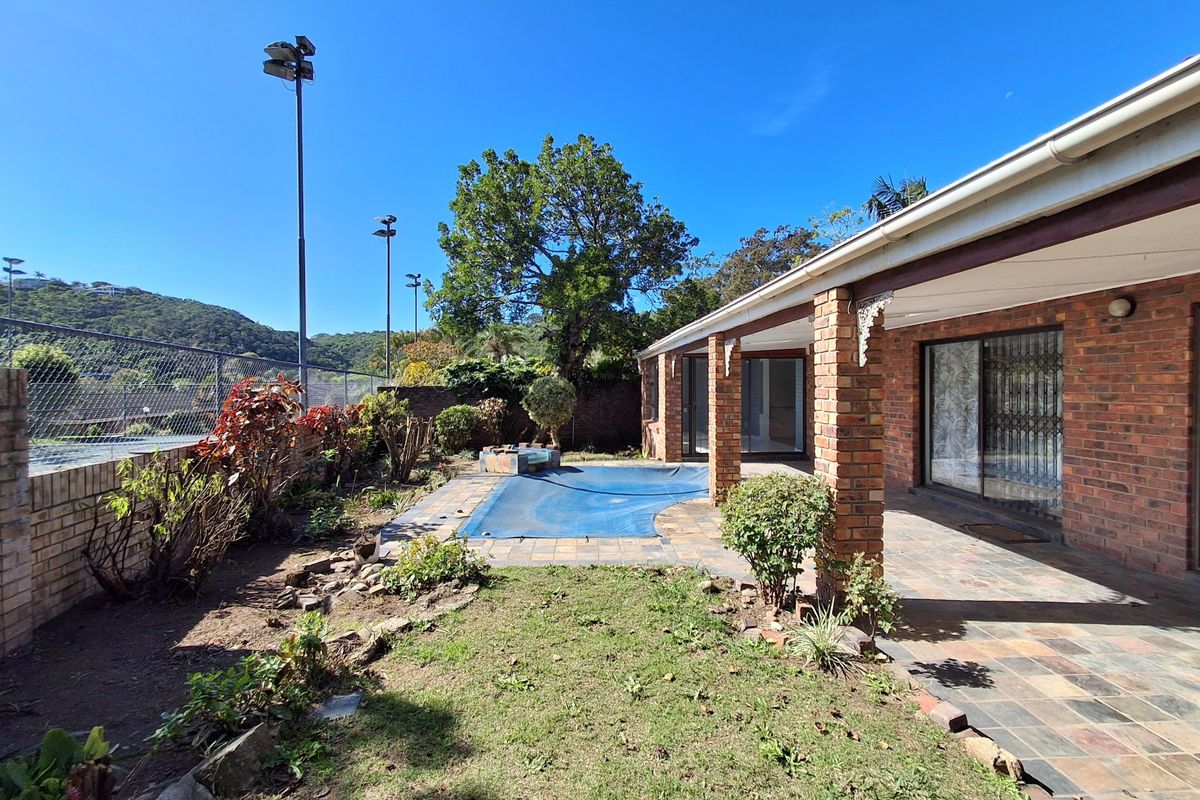 Rear exterior view featuring a covered brick-pillar patio, paved pool area with cover, and adjacent sports court fencing.