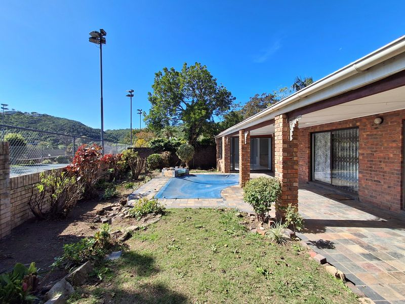 Rear exterior view featuring a covered brick-pillar patio, paved pool area with cover, and adjacent sports court fencing.
