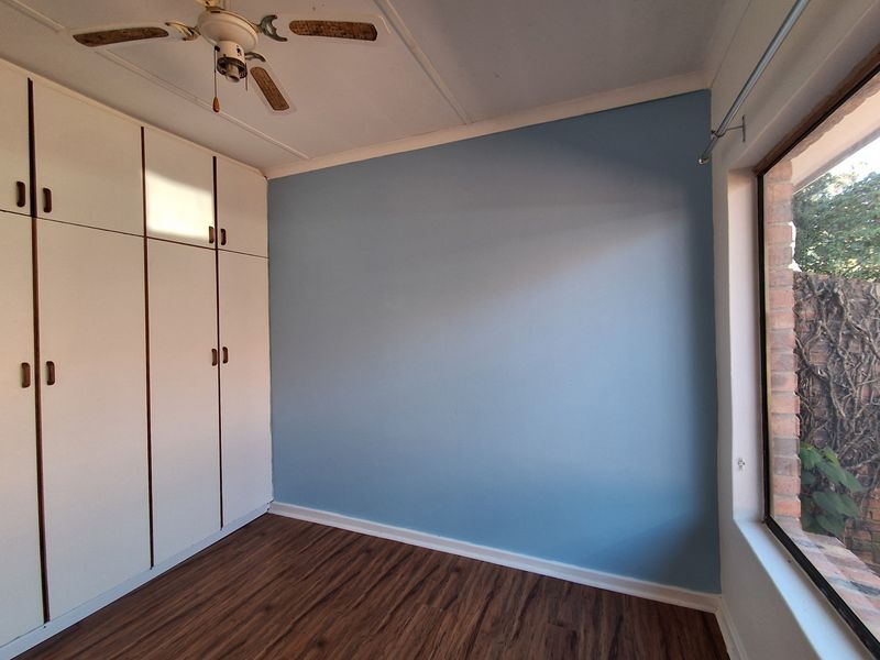 A bright, empty bedroom featuring built-in cupboards, a ceiling fan, and laminate flooring, with a large window overlooking the exterior garden area.