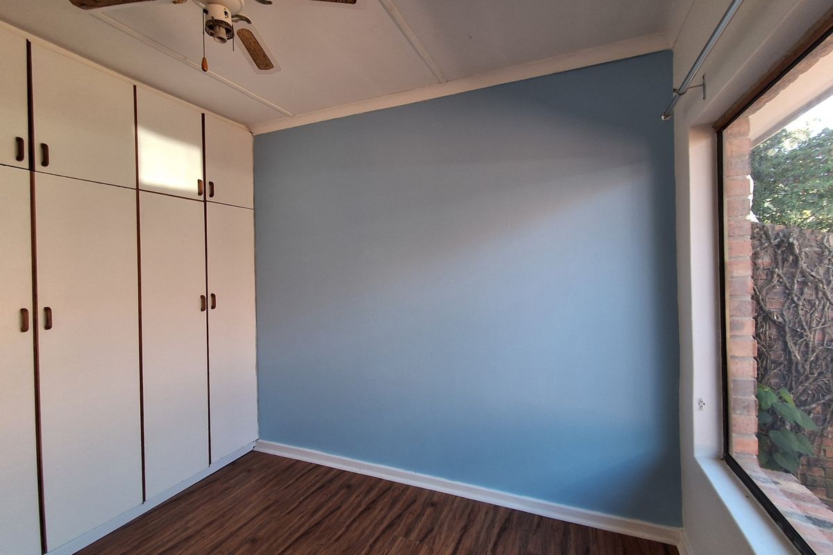 A bright, empty bedroom featuring built-in cupboards, a ceiling fan, and laminate flooring, with a large window overlooking the exterior garden area.
