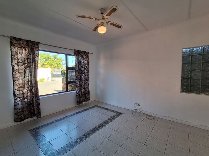 A bright, empty living room featuring tiled flooring with a decorative border, a ceiling fan, and a unique glass block feature window.