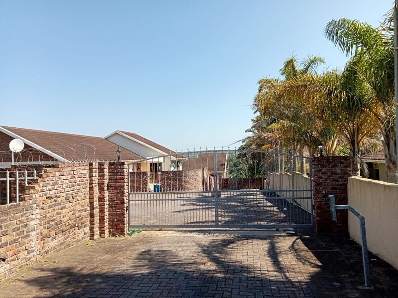 Front entrance view of the cluster property featuring a paved driveway, secure metal gate, and brick boundary wall with security fencing.