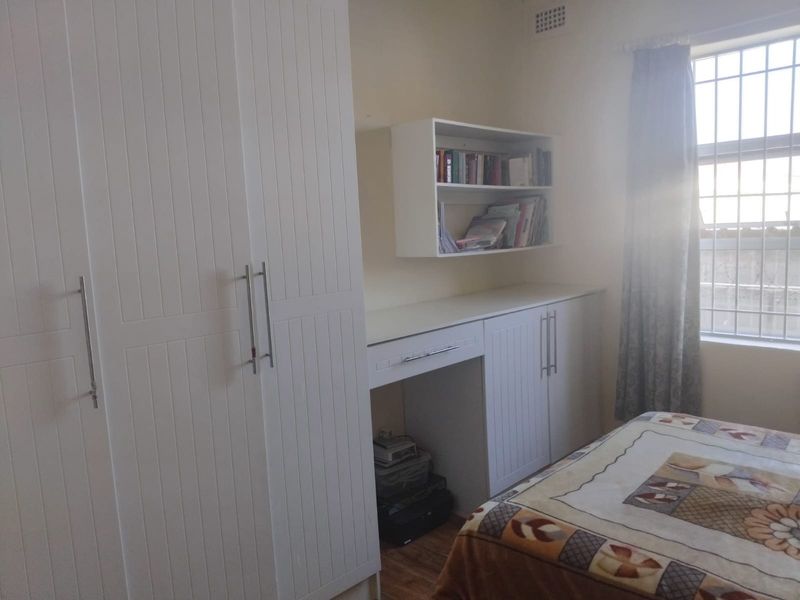 A bedroom featuring white built-in cabinetry, including a wardrobe and a desk with overhead shelving, complemented by a window with security bars.