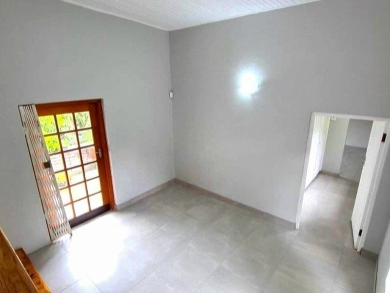 A bright, empty living area featuring neutral tiled flooring, a wooden framed glass door leading outside, and a wooden staircase railing.