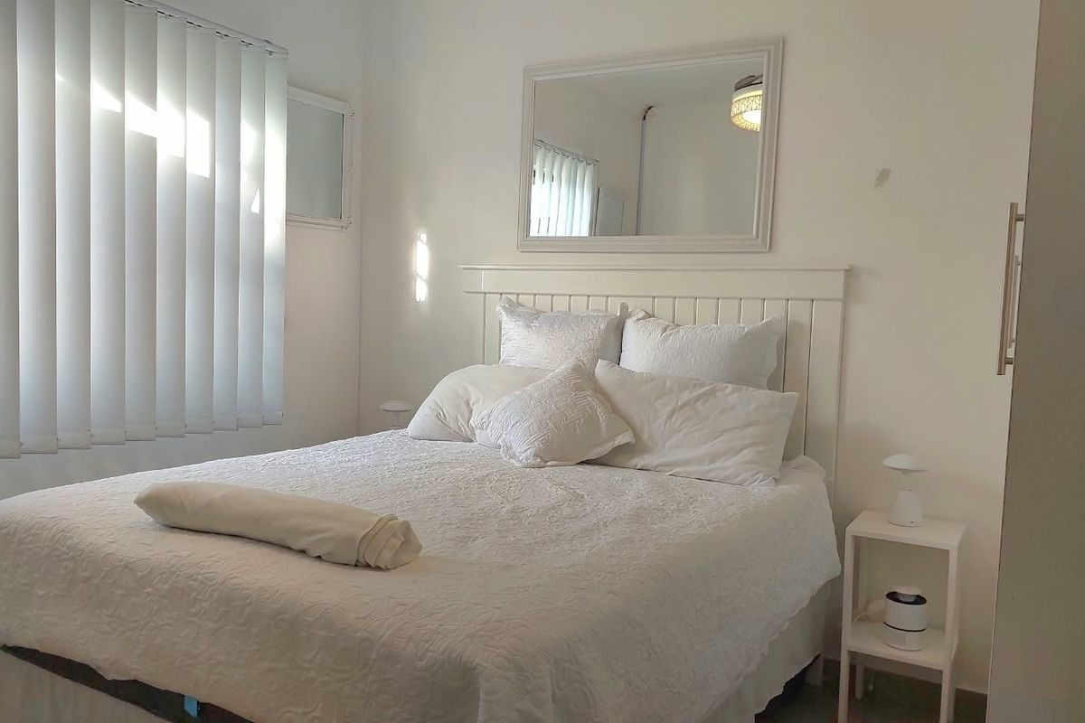 A bright bedroom featuring a built-in headboard, vertical window blinds, and a wall-mounted mirror, offering a clean and neutral living space.