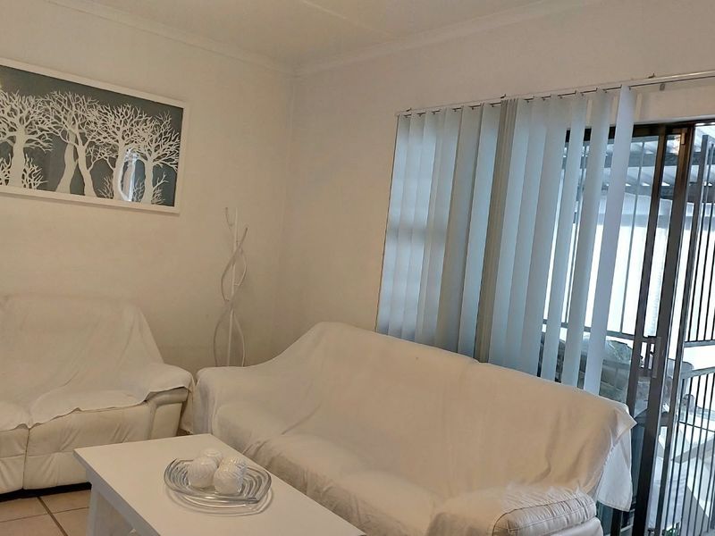 A bright living room featuring tiled flooring, vertical blinds, and a sliding glass door with an integrated security gate leading to an outdoor area.