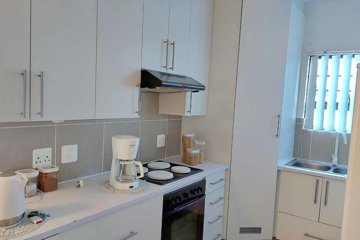 A clean, modern kitchen featuring white cabinetry, a built-in oven with electric hob, extractor fan, and a stainless steel sink with tiled backsplash.