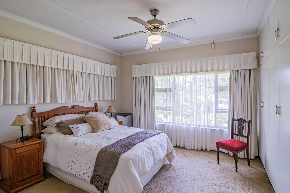 A well-lit bedroom featuring built-in cupboards, a ceiling fan, and large windows with neutral curtains, offering a comfortable and functional living space.