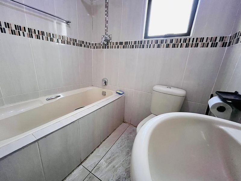 A functional bathroom featuring a built-in bathtub, pedestal basin, and toilet with neutral wall tiling and decorative mosaic accents.