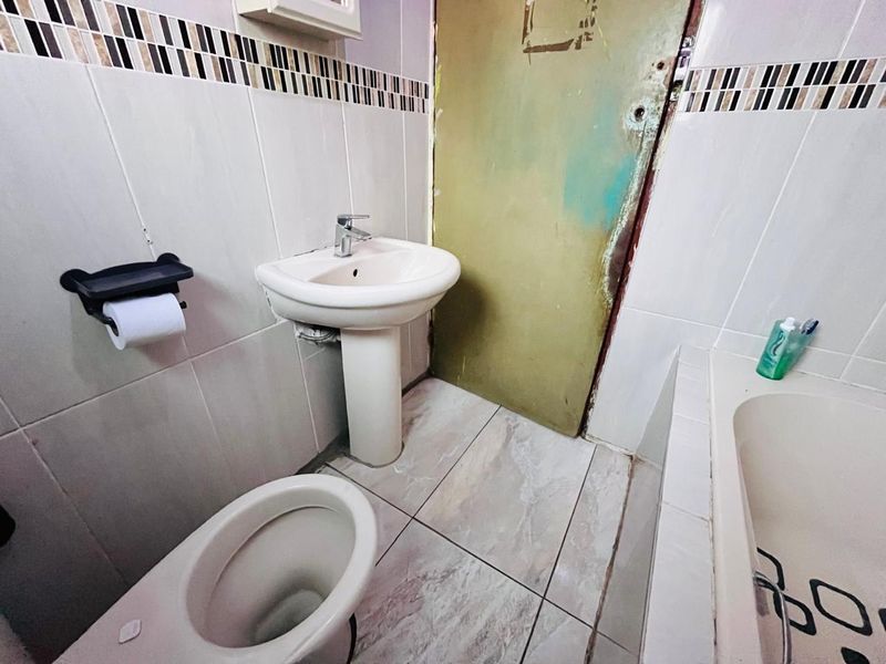 A compact bathroom featuring a pedestal basin, toilet, and bathtub with tiled finishes, requiring cosmetic updates to the door and general maintenance.
