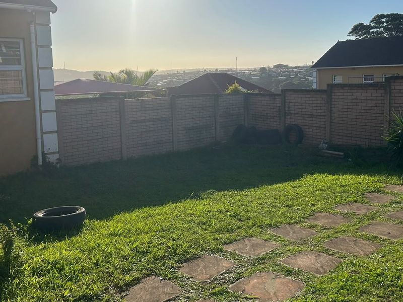 A rear garden area featuring a brick boundary wall, a paved walkway, and a lawn with distant neighborhood views.