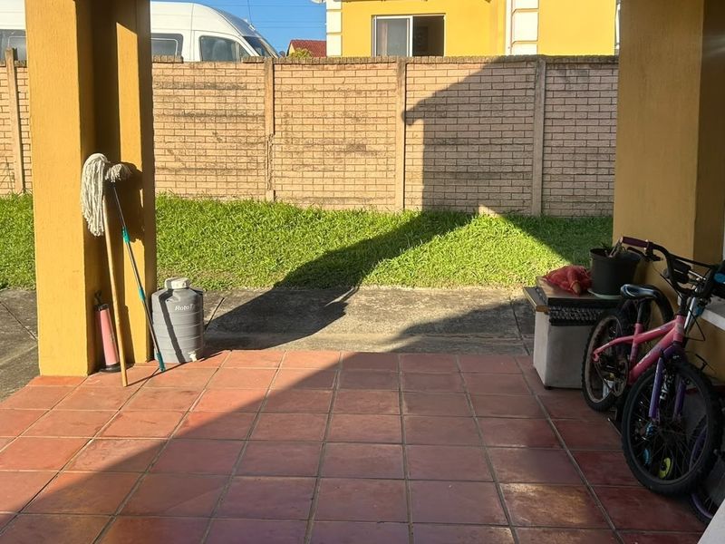 A covered patio area featuring tiled flooring and concrete pillars, overlooking a private garden space with a brick boundary wall.