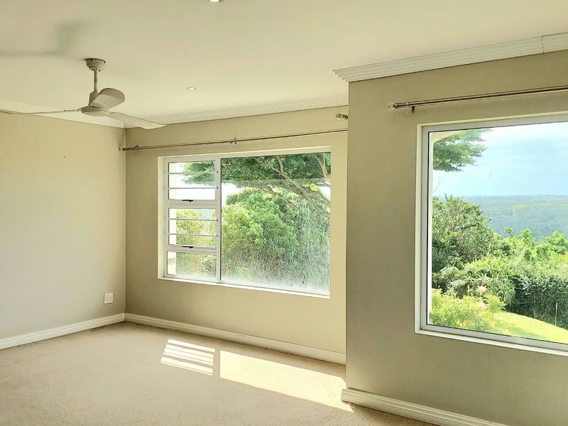 A bright, empty bedroom featuring neutral walls, carpeted flooring, recessed lighting, a ceiling fan, and large windows offering scenic views.