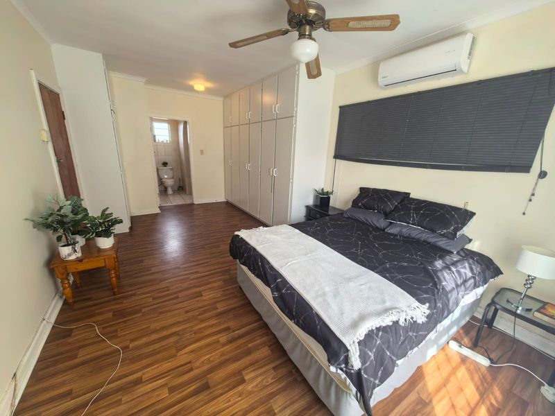 Spacious bedroom featuring laminate flooring, extensive built-in cupboards, a ceiling fan, and a wall-mounted air conditioning unit, with direct access to an en-suite bathroom.