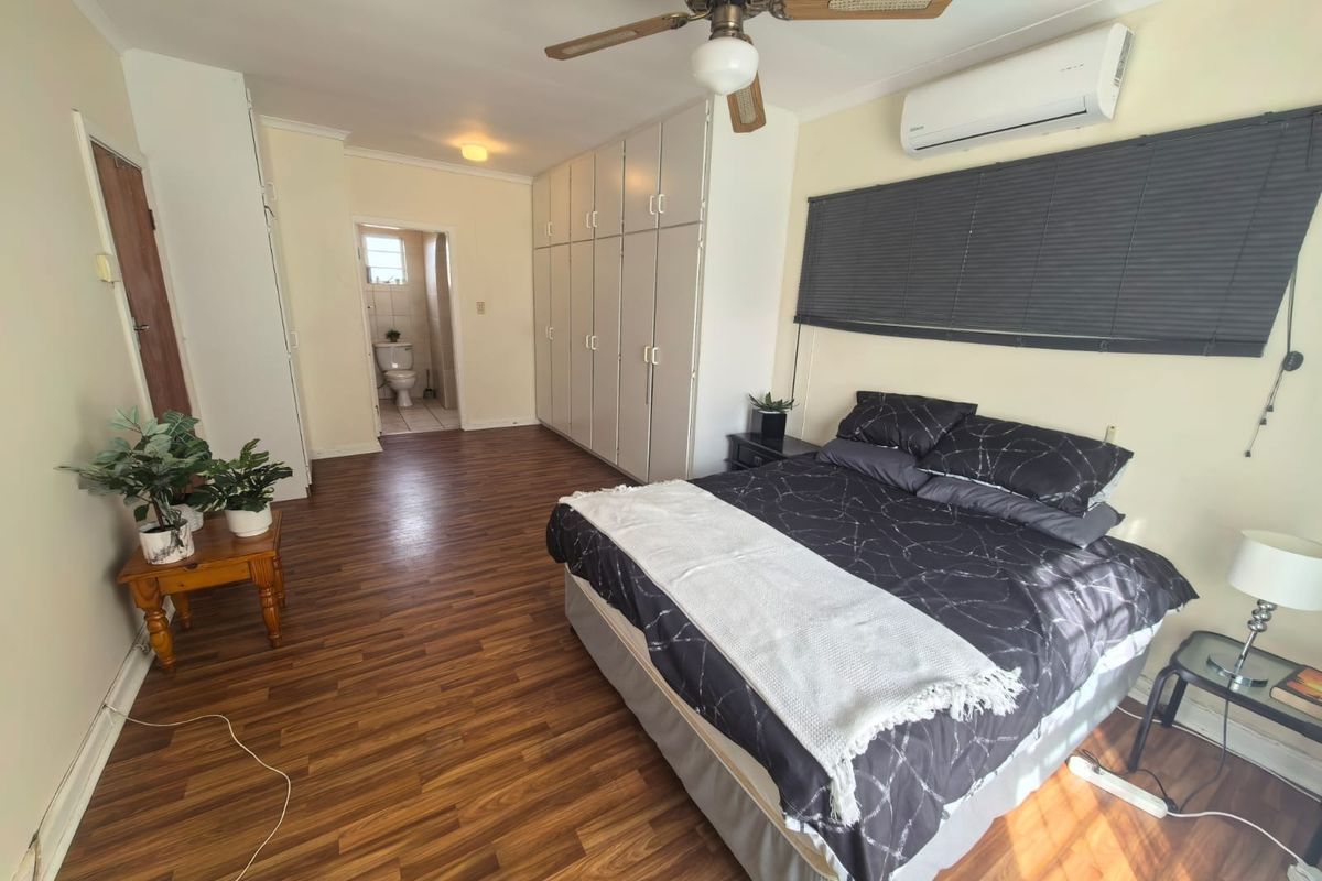 Spacious bedroom featuring laminate flooring, extensive built-in cupboards, a ceiling fan, and a wall-mounted air conditioning unit, with direct access to an en-suite bathroom.