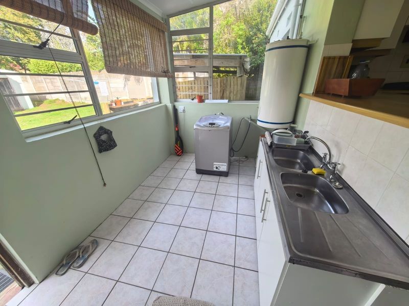 Functional laundry area featuring a double stainless steel sink, tiled flooring, and an integrated electric geyser with ample natural light from louvered windows.
