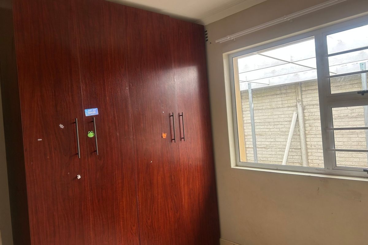 A bedroom featuring built-in wooden cupboards and an aluminium window with security bars, offering a functional and neutral space.